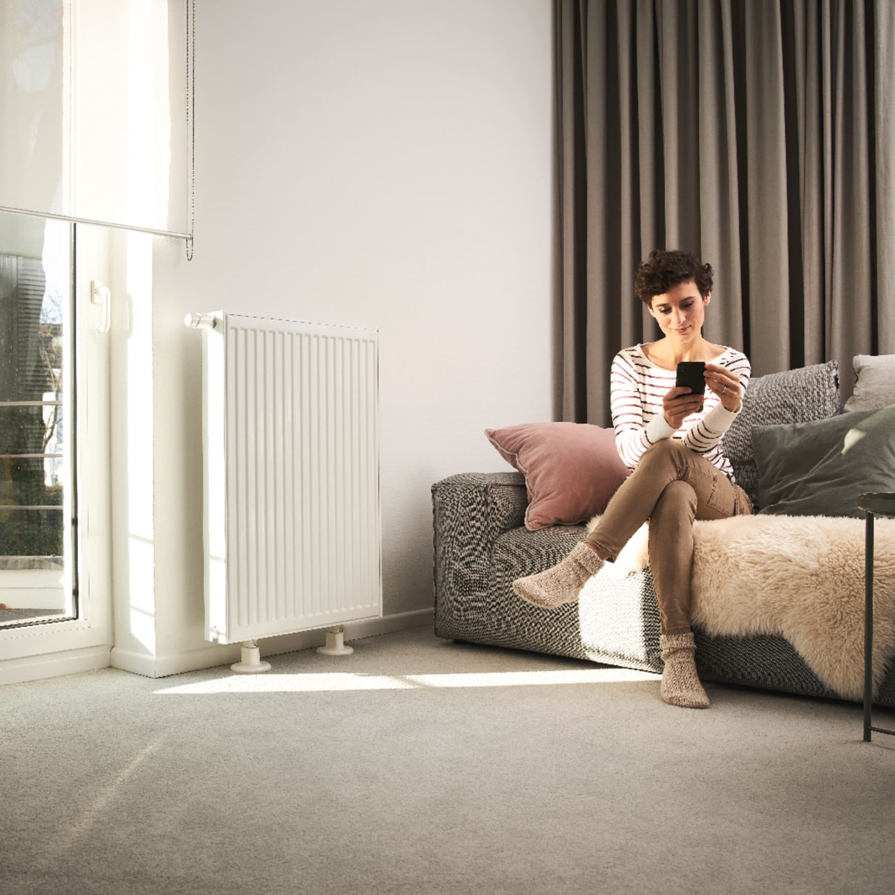 Une femme est assise sur un canapé et utilise un smartphone pour régler son chauffage. Le canapé se trouve devant une fenêtre et à côté d'un radiateur | Vaillant