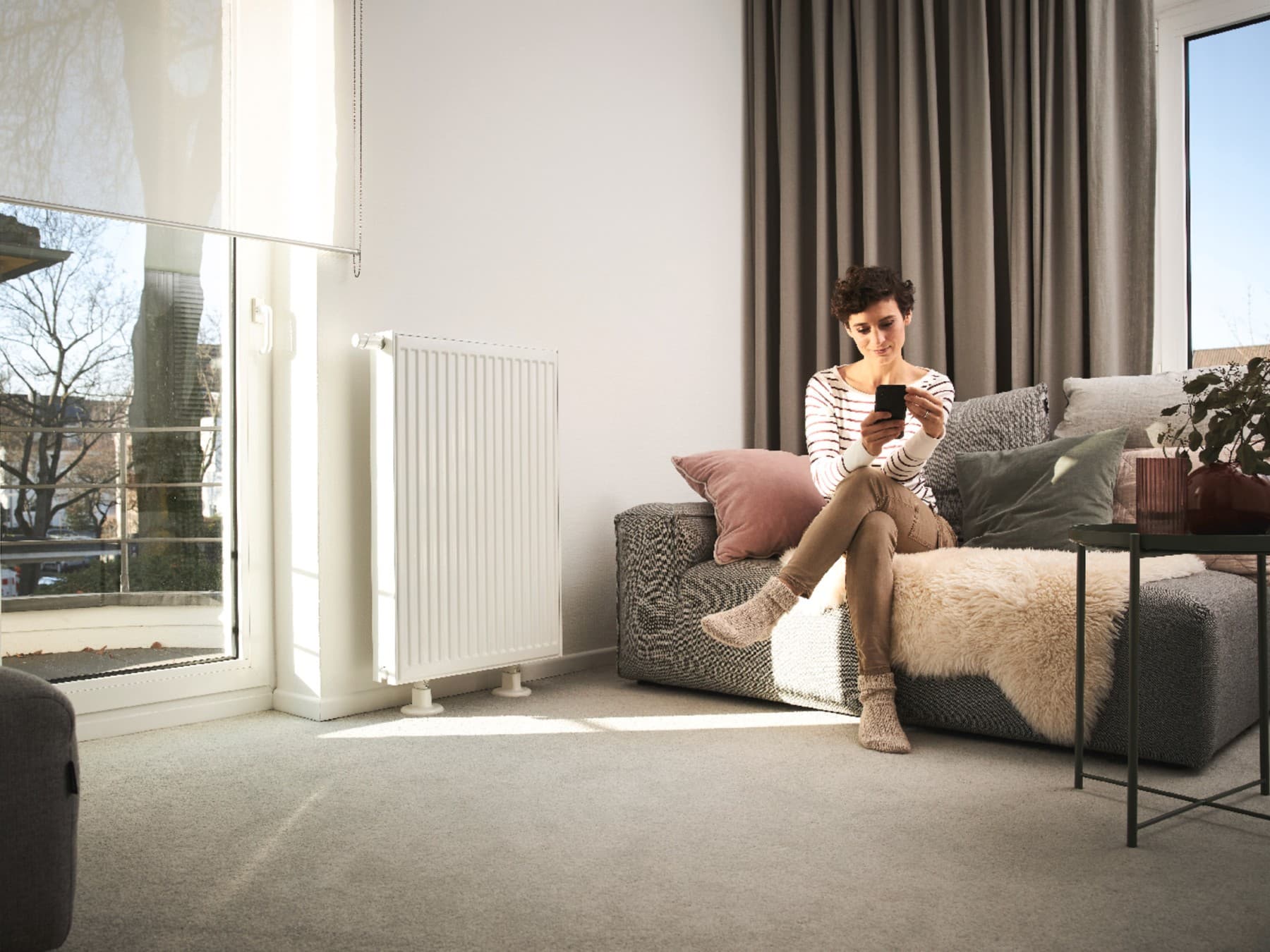 Une femme est assise sur un canapé et utilise un smartphone. Le canapé se trouve devant une fenêtre et à côté d'un radiateur | Vaillant