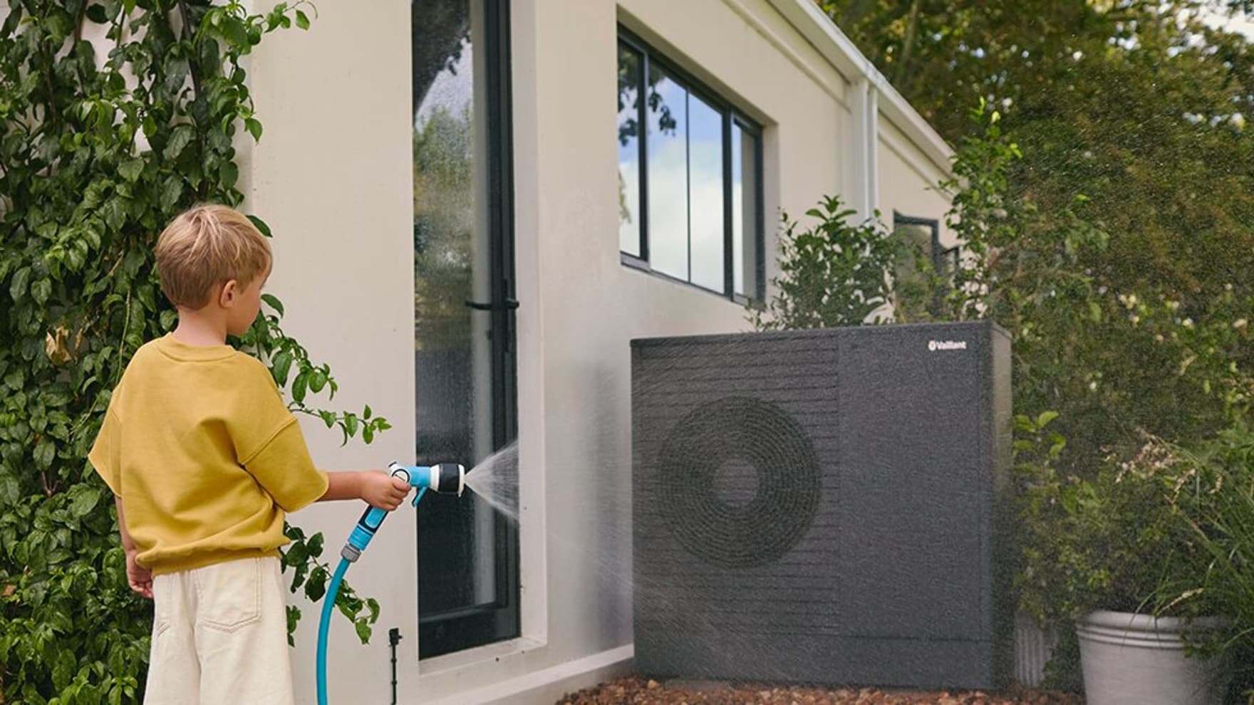 Un garçon arrose la pompe à chaleur avec un tuyau d'arrosage | Vaillant