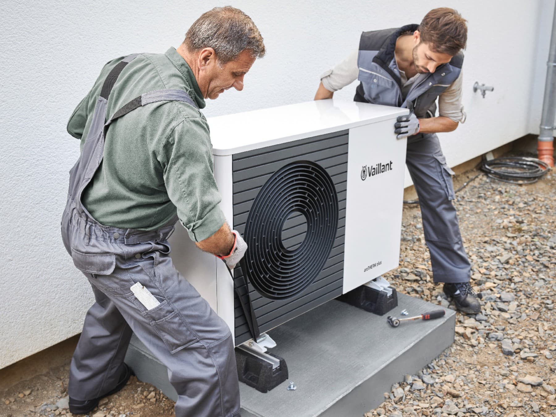 Deux installateurs certifiés installent une unité extérieure de pompe à chaleur dans une cour | Vaillant