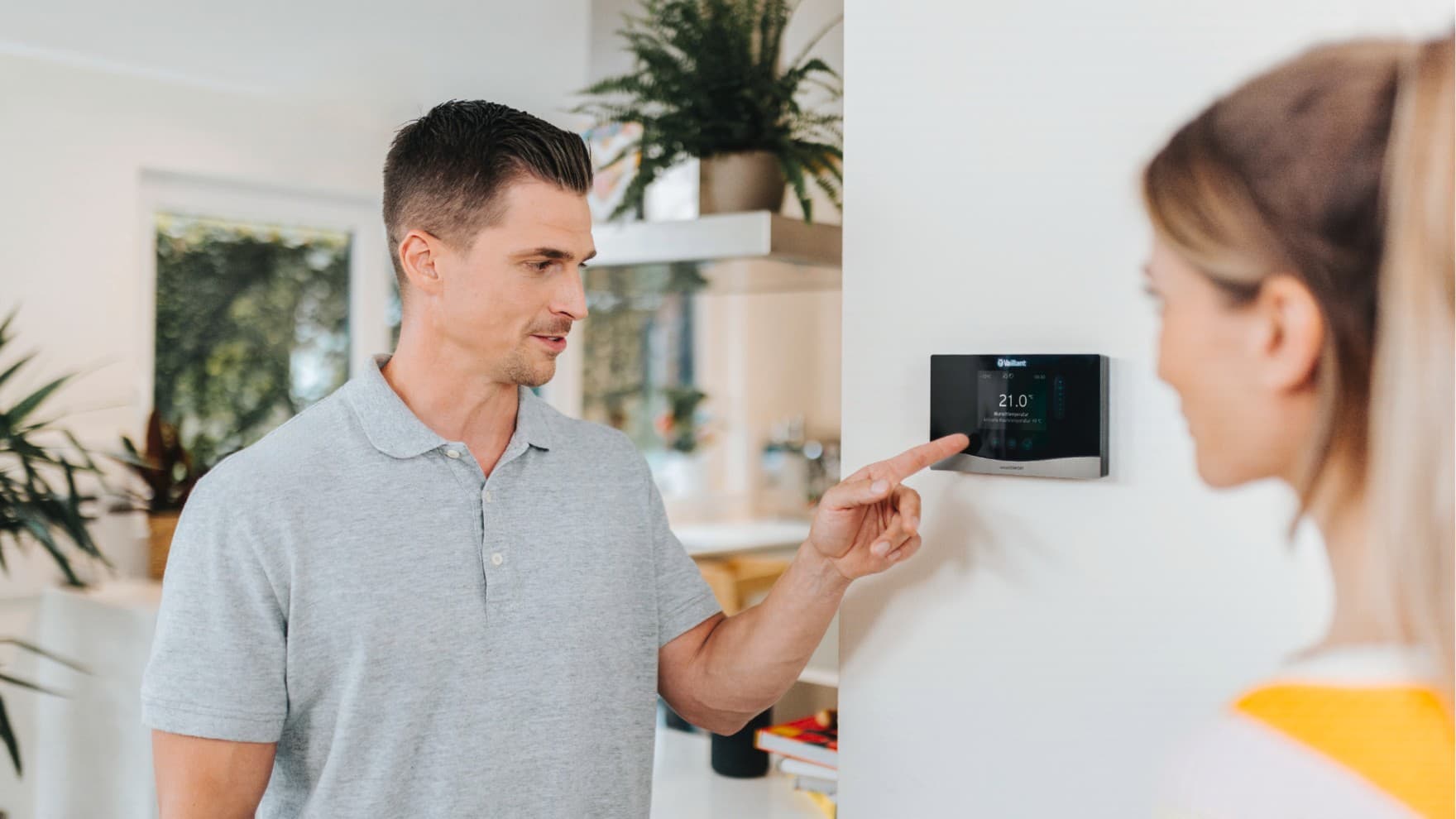 Un homme est au premier plan, la main posée sur un thermostat fixé à un mur bleu | Vaillant