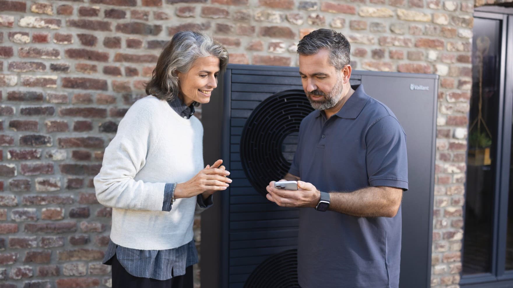 Installateur et une propriétaire regardant un smartphone devant une pompe à chaleur | Vaillant