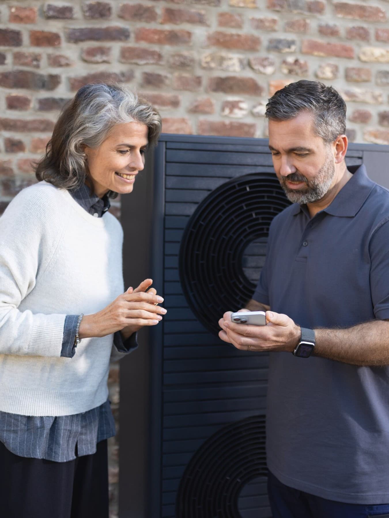 Installateur et une propriétaire regardant un smartphone devant une pompe à chaleur | Vaillant