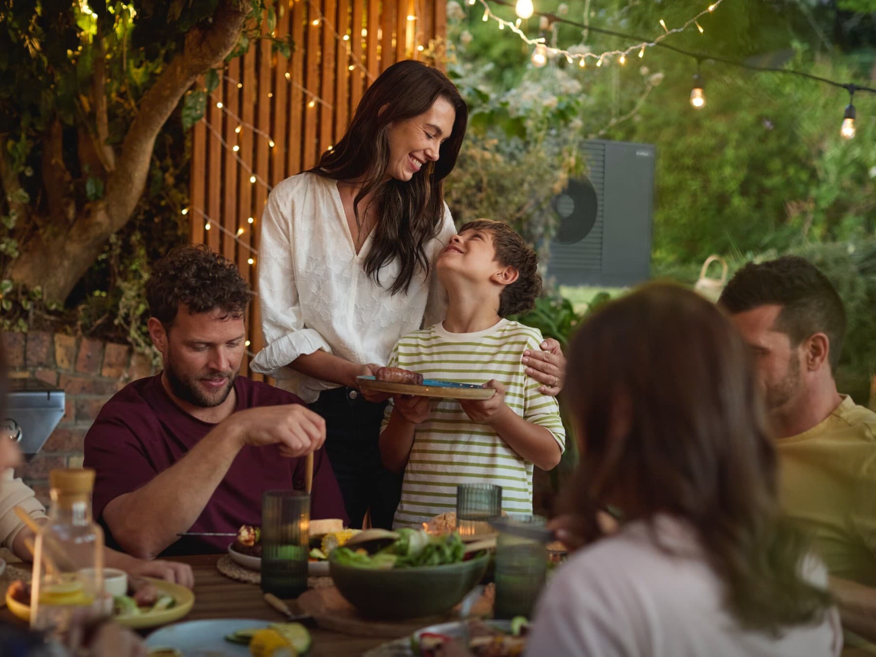 Une famille profitant d'un barbecue dans le jardin avec des amis. Une unité extérieure de pompe à chaleur dans le fond.