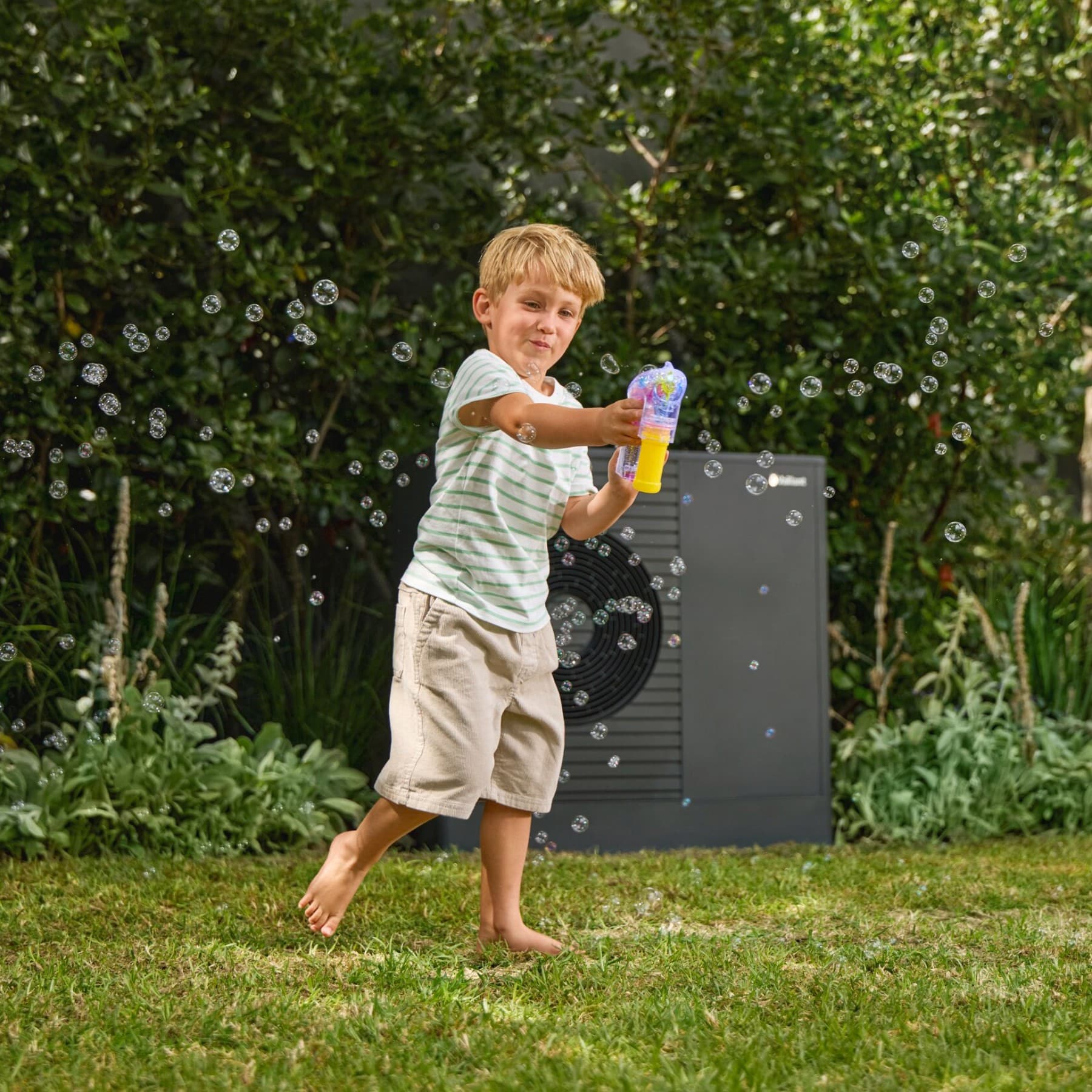 Garçon qui joue à faire des bulles de savon dans son jardin avec une pompe à chaleur au fond | Vaillant