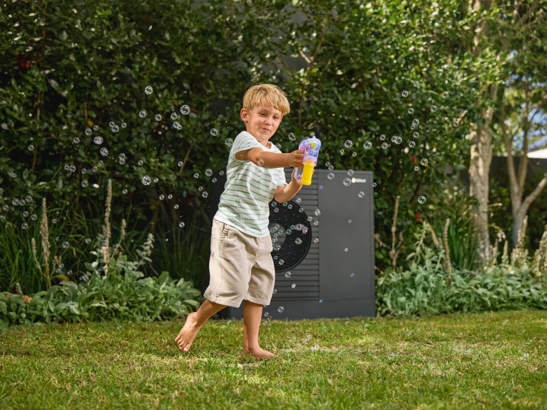 Garçon qui joue à faire des bulles de savon dans son jardin avec une pompe à chaleur au fond | Vaillant