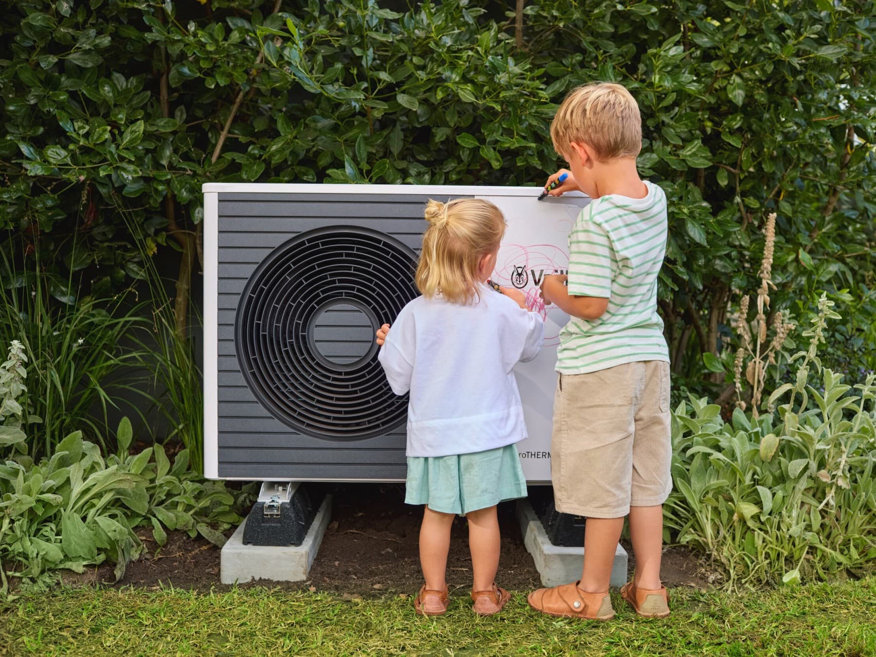 Deux enfants dessinent sur une unité de pompe à chaleur dans un jardin| Vaillant